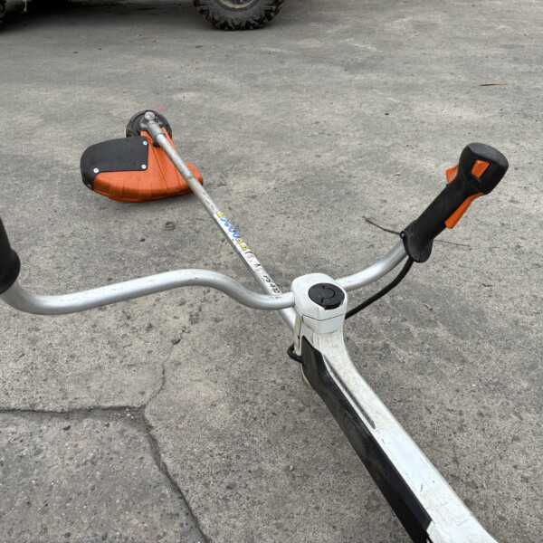 Lawn trimmer with an orange guard lying on a concrete driveway, handle in the foreground.