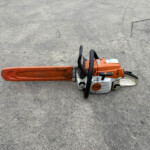 Orange STIHL chainsaw resting on rough gray asphalt, bar extending to the left and black handles visible.