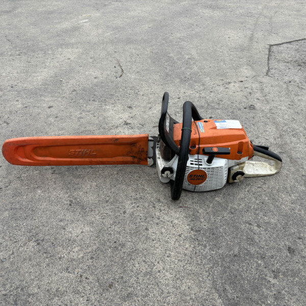 Orange STIHL chainsaw resting on rough gray asphalt, bar extending to the left and black handles visible.