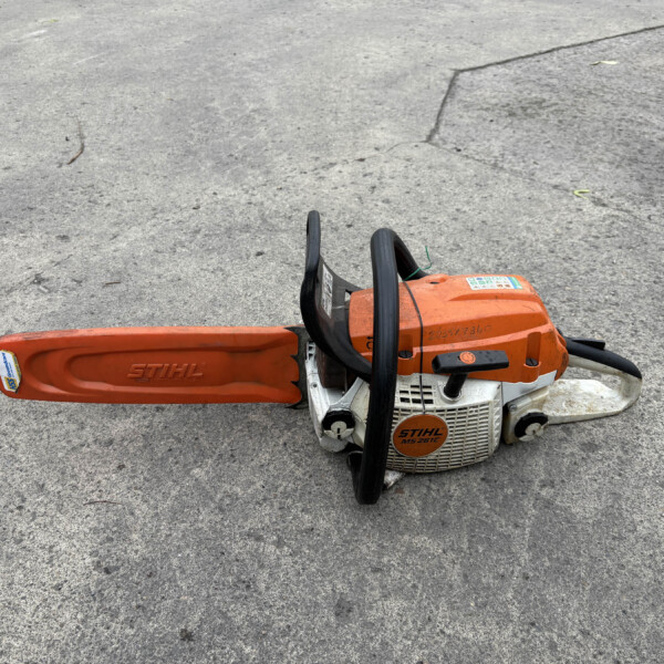 Orange STIHL chainsaw lying on a gray asphalt surface, bar extending to the left.