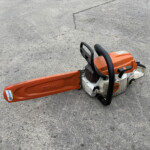 Orange STIHL chainsaw lying on a rough concrete surface, bar and handles visible.