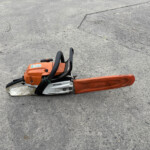 Orange STIHL chainsaw resting on a rough concrete surface, seen from the side with the bar partially visible at the lower right.