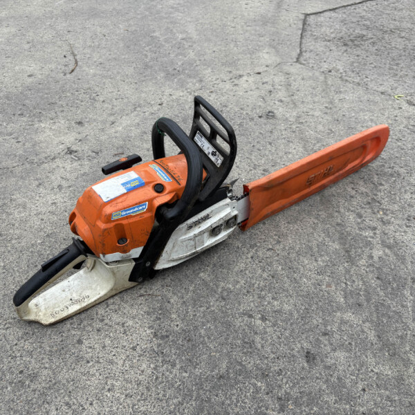 Orange chainsaw resting on cracked gray asphalt, viewed from above on the groundjoist surface.