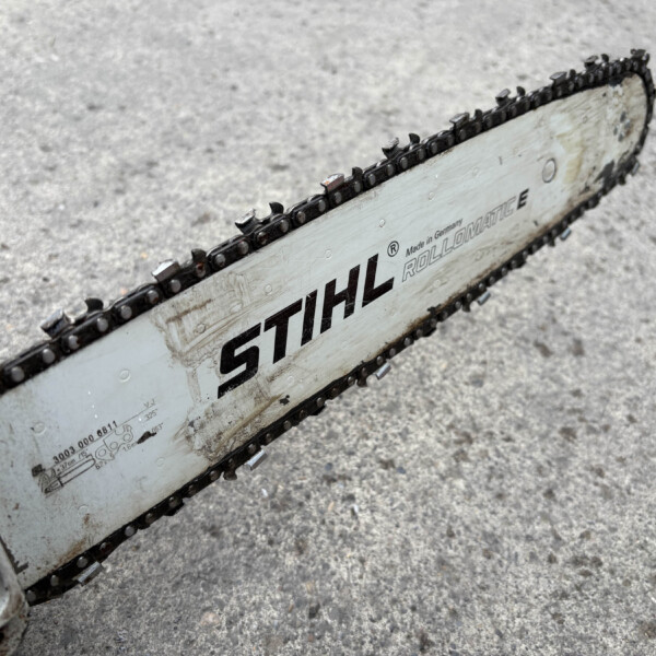 Close-up of a chainsaw bar and chain resting on a rough concrete surface, showing the STIH L (STIHL) branding and worn edges.