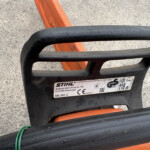 Close-up of a black chainsaw bar with an orange handle, lying on a rough concrete surface.