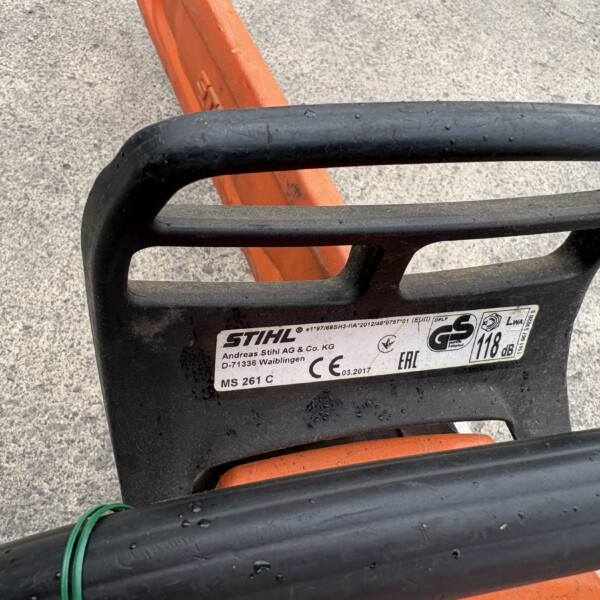 Close-up of a black chainsaw bar with an orange handle, lying on a rough concrete surface.
