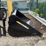Black Skid-Steer bucket attachment labeled STRIMECH resting on gravel beside a green metal guardrail, with part of a tractor on the left.