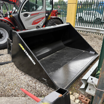 Manitou telehandler with a large black bucket attachment parked on a gravel lot beside a green fence.
