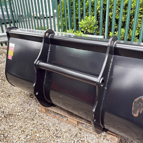 Large black metal construction bucket resting on a wooden pallet outdoors, with a green fence and shrubs in the background.