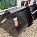 Black metal heavy-duty bucket attachment with warning labels resting on gravel beside a green fence.