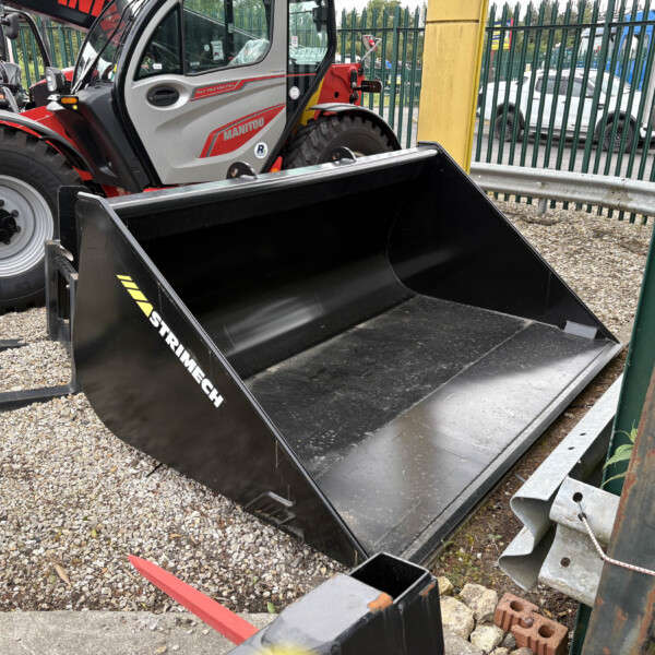 Manitou telehandler with a large black bucket attachment parked on a gravel lot beside a green fence.
