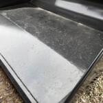 Close-up of a black metal trailer floor with grey aluminum edge panels and a gravel surface along the side outside the trailer.