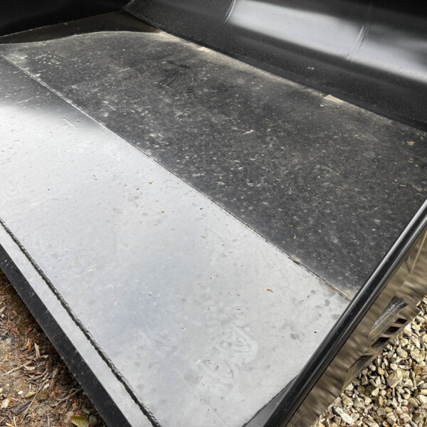 Close-up of a black metal trailer floor with grey aluminum edge panels and a gravel surface along the side outside the trailer.