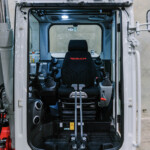 Open cab of a Takeuchi construction machine with an operator seat, joysticks, and control panel visible at the front.