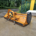 Orange heavy-duty ground-maintenance machine on a wet gravel surface, with a green fence and trees in the background.