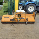 Front view of an orange Teagle mower attachment on a concrete surface with blue tractors in the background.