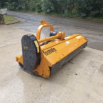 Orange industrial mower attachment on a concrete surface, with a wet road and trees in the rainy background