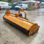 Orange road cutter/soil grinder on a wet concrete surface in a construction yard, with white vans and branded flags visible in the background.