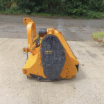 Orange stump grinder/wood chipper with a large toothed cutting wheel on a concrete area outdoors, with greenery in the background.