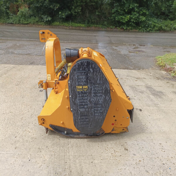 Orange stump grinder/wood chipper with a large toothed cutting wheel on a concrete area outdoors, with greenery in the background.