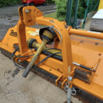 Close-up of an orange hydraulic-powered mower attachment with hoses and rotating shaft on a concrete surface outdoors