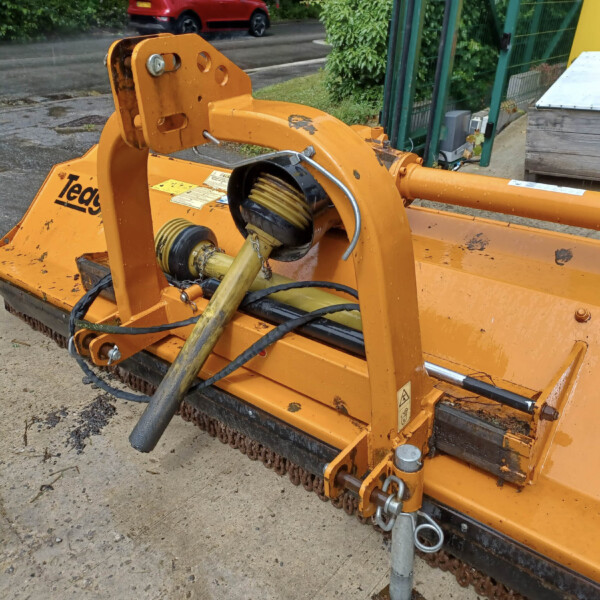 Close-up of an orange hydraulic-powered mower attachment with hoses and rotating shaft on a concrete surface outdoors