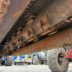 Close-up of a rusted agricultural machine underside showing metal gears, chains, and track links in a dusty workshop setting