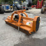 Orange agricultural implement with chain-driven roller in the foreground, parked in an equipment yard with blue tractors and red machinery in the background.
