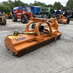 Orange Teagle farm mower with metal tines in a gravel lot among other agricultural machines.