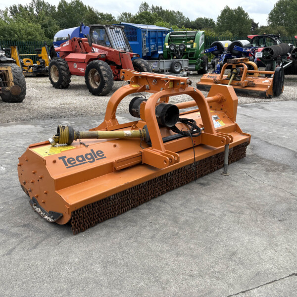 Orange Teagle farm mower with metal tines in a gravel lot among other agricultural machines.