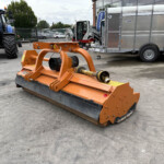 Orange heavy-duty hydraulic attachment on the pavement with a blue tractor and a large silver trailer in the background.