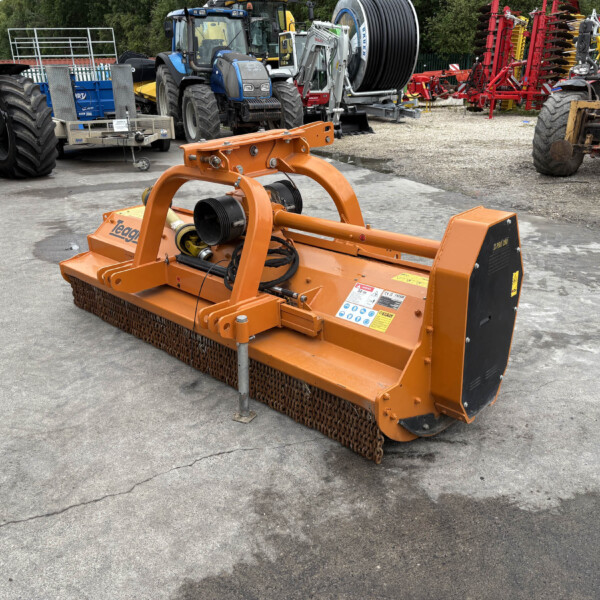 Orange agricultural implement with chain-driven roller in the foreground, parked in an equipment yard with blue tractors and red machinery in the background.