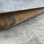 Close-up of a rusted metal roller beneath a gray metal sheet on a rough concrete floor.
