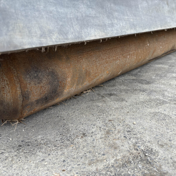 Close-up of a rusted metal roller beneath a gray metal sheet on a rough concrete floor.