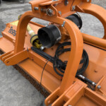 Orange industrial attachment with pulleys, belts, and hydraulic lines mounted on a low-profile machine, outdoors on concrete surface