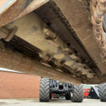 Close-up of a rusty heavy machinery undercarriage with metal tracks and chains showing wear and dirt, under a large orange vehicle.