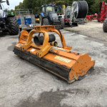Orange heavy machinery attachment in foreground on a concrete lot, with blue tractors and large reel-spool equipment in the background on a workyard.
