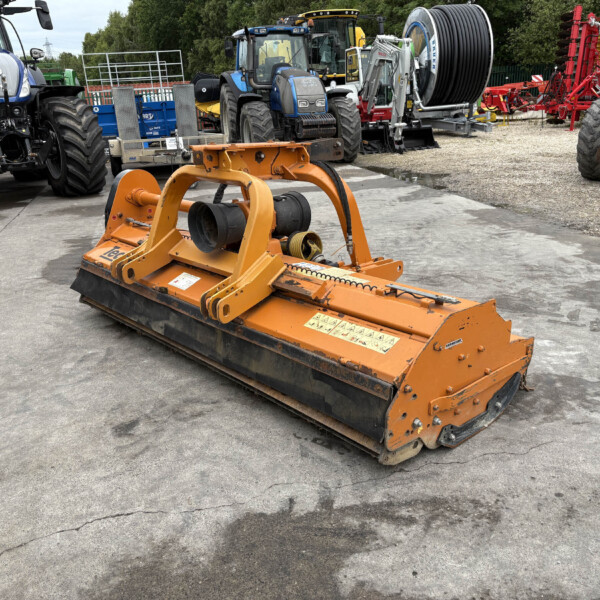 Orange heavy machinery attachment in foreground on a concrete lot, with blue tractors and large reel-spool equipment in the background on a workyard.