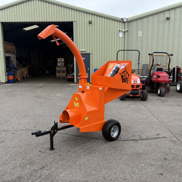 Orange industrial wood chipper/extender in the foreground outside a green corrugated-metal warehouse, with pallets and a worker visible inside the open door and red tractors lined up to the right.