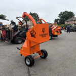 Orange Timberwolf PTO/100G log splitter on wheels in an outdoor yard with stacked pallets and other machinery in the background, logo visible on the side.