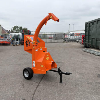 Orange industrial machine with a curved, reach arm in a yard, surrounded by other equipment and a tall green dumpster on the right.