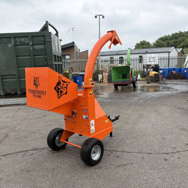 Orange Timberwolf wood chipper in a yard, with a large green dumpster on the left and industrial buildings behind, under a cloudy sky.