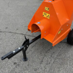 Orange metal industrial frame with warning labels, attached to a black hitch and small wheeled base on asphalt