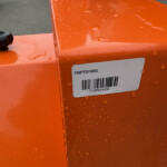Close-up of bright orange metal surface with a black knob, water droplets, and a barcode label.