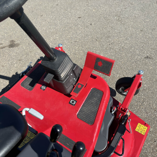 Red pedal-powered toy tractor viewed from above, showing pedals, black seat, and steering lever on a paved surface
