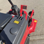 Red tractor control area with three black levers, pedals, and a yellow caution label on the console surface, outdoors on asphalt.