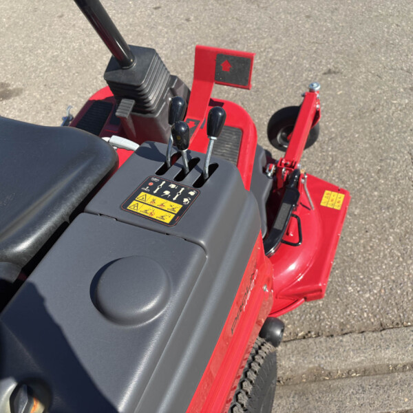Red tractor control area with three black levers, pedals, and a yellow caution label on the console surface, outdoors on asphalt.