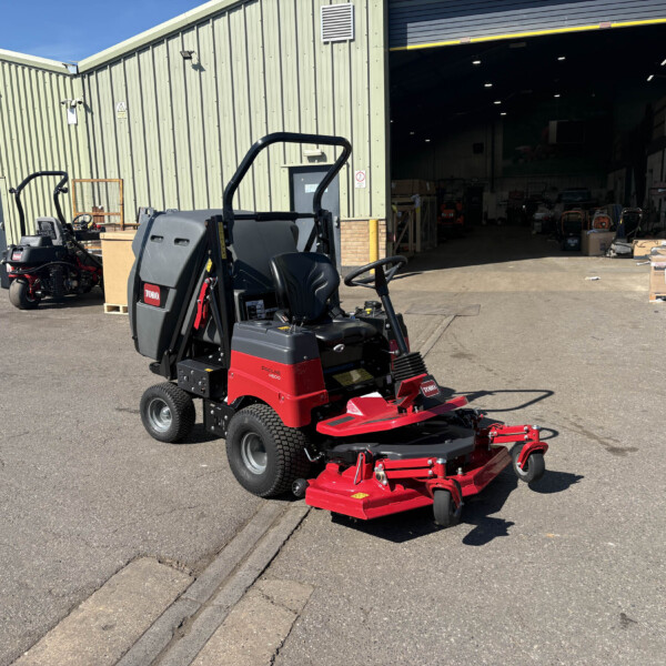 Red and black riding lawn mower in an outdoor industrial yard, with a large metal warehouse behind and scattered boxes and equipment nearby