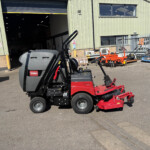 Red and gray Toro ProLine H6000 ride-on mower with a large mower deck attached, parked outside a green corrugated metal warehouse with a wide open bay door behind it.
