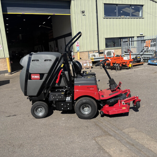 Red and gray Toro ProLine H6000 ride-on mower with a large mower deck attached, parked outside a green corrugated metal warehouse with a wide open bay door behind it.